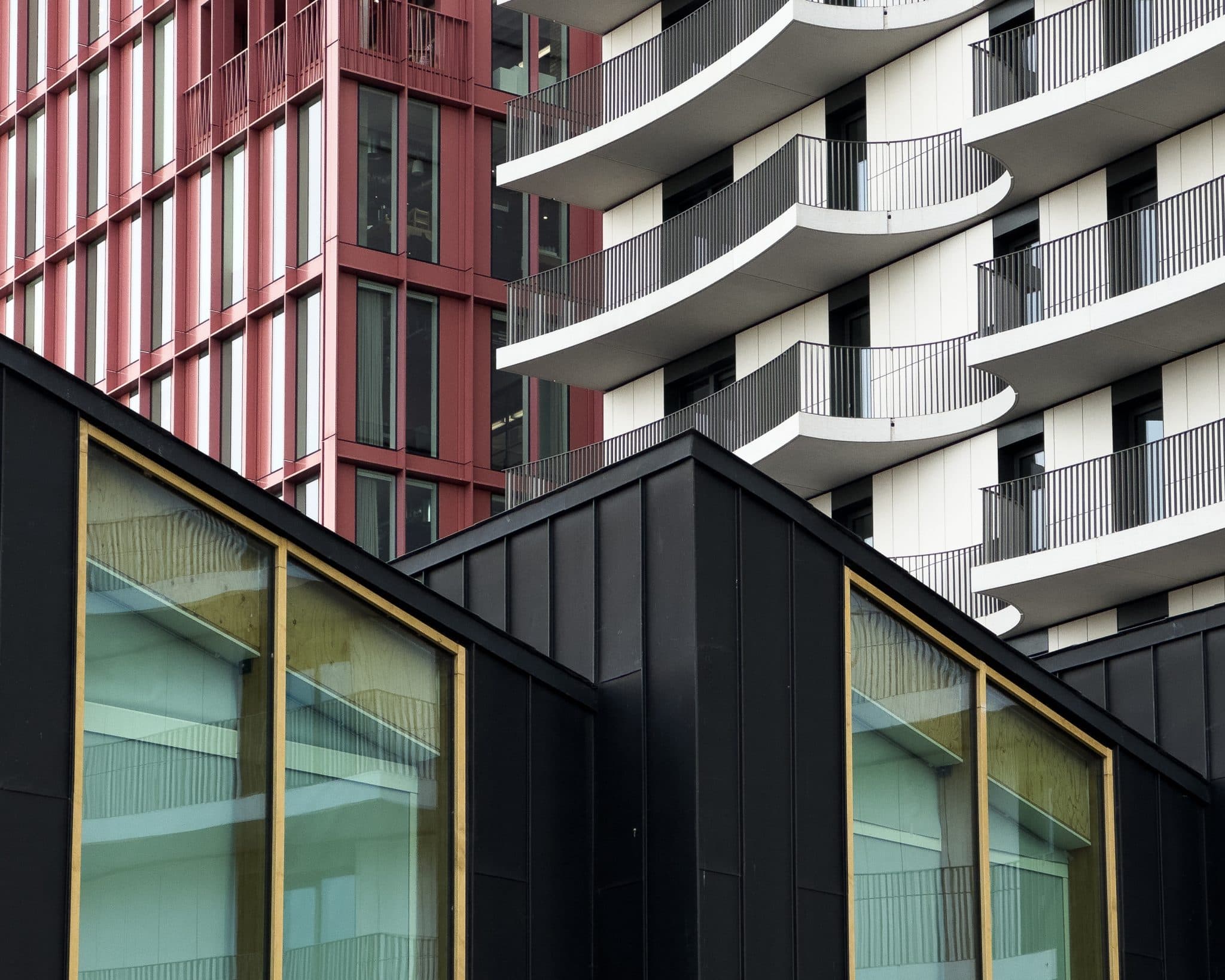 Dettaglio ravvicinato di edifici residenziali contemporanei sovrapposti: in basso un volume con rivestimento metallico nero a doghe verticali e grandi vetrate con telai in legno chiaro; dietro, una palazzina con facciata a montanti rosso mattone e, a destra, un edificio bianco con balconi dalle linee curve e ringhiere verticali nere.