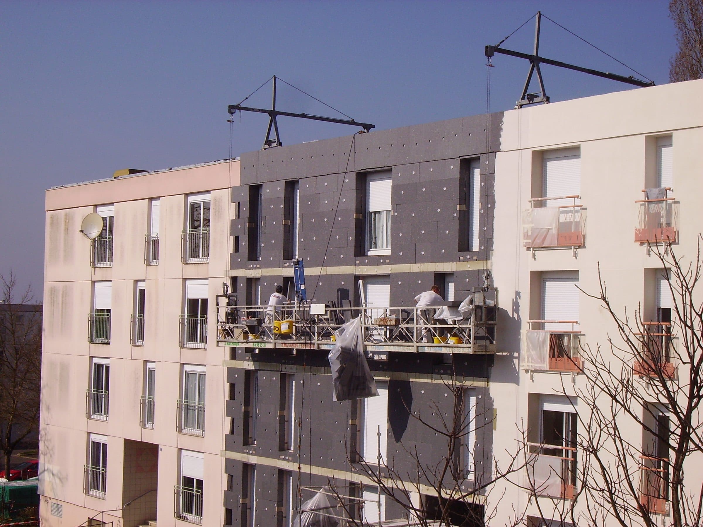 Facciata di un condominio durante la posa del cappotto termico: sulla porzione centrale sono già fissati i pannelli isolanti grigio antracite con i tasselli a fungo bianchi a vista, mentre le due porzioni laterali mostrano ancora l'intonaco vecchio color sabbia. Due operai in tuta bianca lavorano da un ponteggio sospeso (cestello su funi) agganciato a bracci metallici sul tetto. Cielo azzurro terso, luce diurna radente, rami spogli in primo piano a destra.