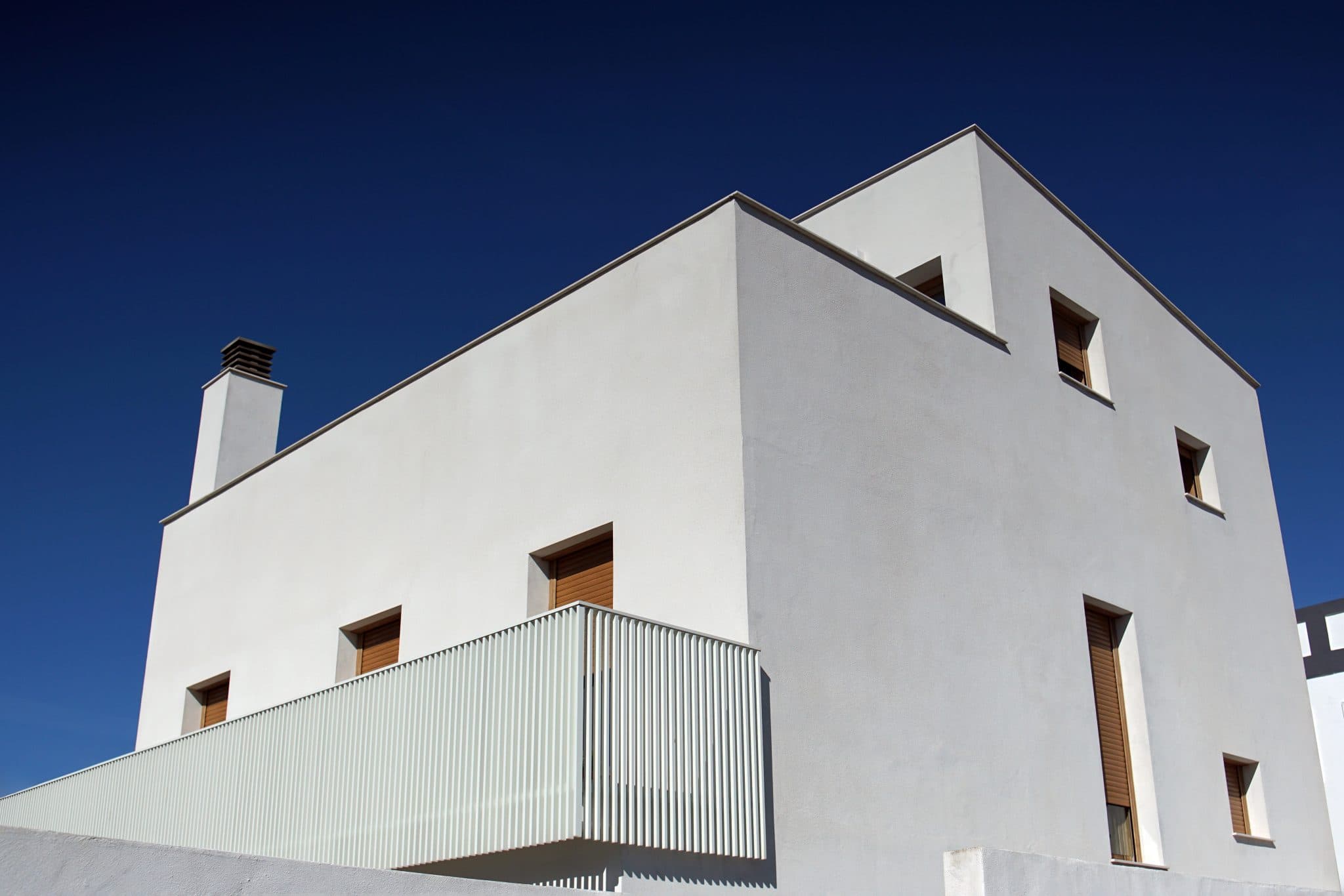 Casa moderna a volumi squadrati con intonaco bianco, vista d'angolo dal basso contro un cielo azzurro intenso; finestre strette con avvolgibili in legno chiaro, balcone rivestito da lamelle metalliche verticali chiaro e un comignolo squadrato sulla sinistra. Luce solare radente che stacca nettamente i due prospetti.