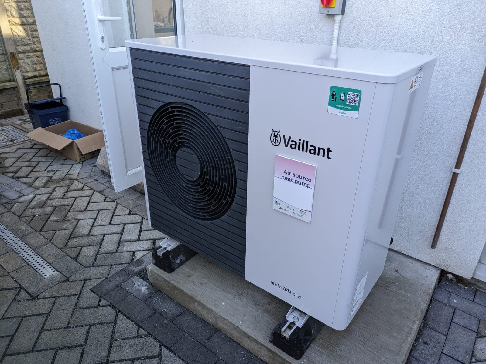 Pompa di calore aria-acqua monoblocco bianca installata all'esterno, appoggiata su due supporti antivibranti a terra e addossata alla parete intonacata chiara di una casa a schiera; in evidenza la grande griglia circolare del ventilatore sul lato scuro dell'unità, la pavimentazione in autobloccanti a spina di pesce e la canalina di scarico, con la porta d'ingresso bianca sullo sfondo e luce naturale diurna
