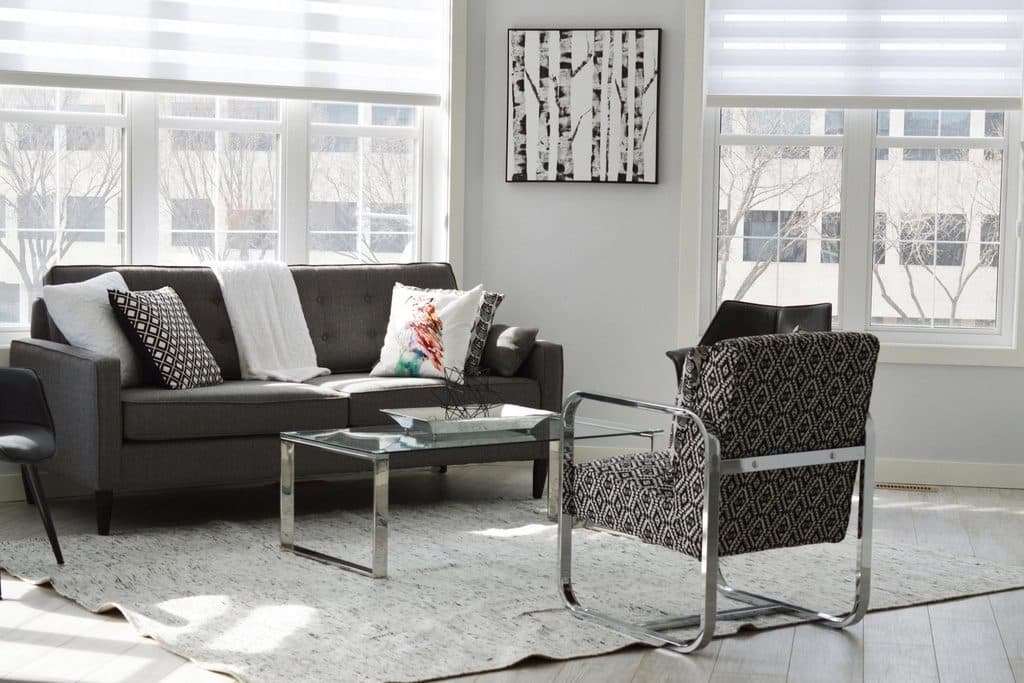 Salotto d'angolo inondato di luce naturale da due grandi finestre con tende a rullo semitrasparenti: divano grigio scuro con cuscini bianchi e a rombi bianco/nero e un plaid bianco, tavolino basso in vetro e acciaio cromato, poltroncina con struttura cromata e tessuto a motivo geometrico bianco e nero, tappeto chiaro irregolare su parquet grigio chiaro, quadro con betulle in bianco e nero alla parete; fuori dalle finestre alberi spogli e la facciata di un edificio.