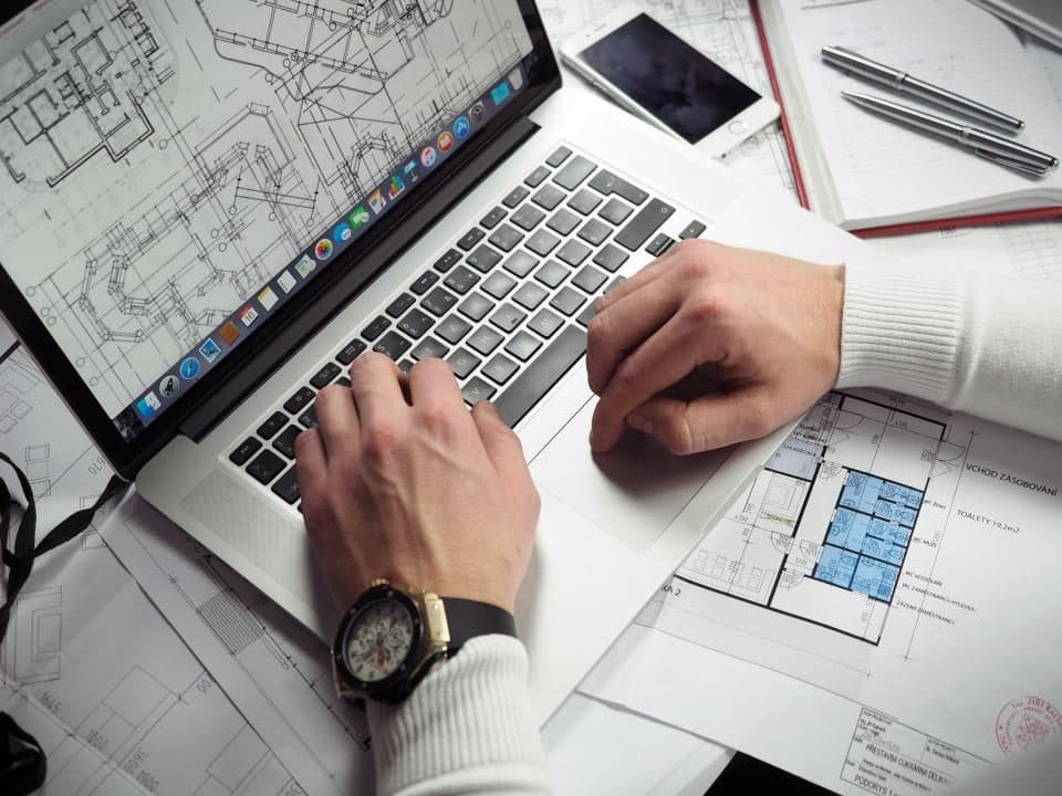 Ripresa dall'alto di una scrivania bianca: due mani maschili con maglione bianco e orologio da polso lavorano su un laptop che mostra a schermo un disegno CAD di pianta architettonica; intorno sono sparse planimetrie stampate in bianco e nero con quote e locali indicati, un telefono, un taccuino aperto e due penne d'argento.